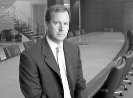 Black-and-white photo of a man in a suit seated at a curved conference table in a modern office setting.